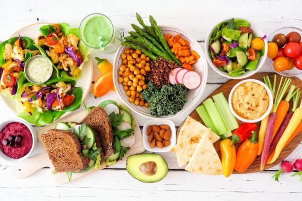 White table with healthy plant-based foods including Buddha bowl, lettuce wraps, colorful vegetables, grains, legumes, and dips